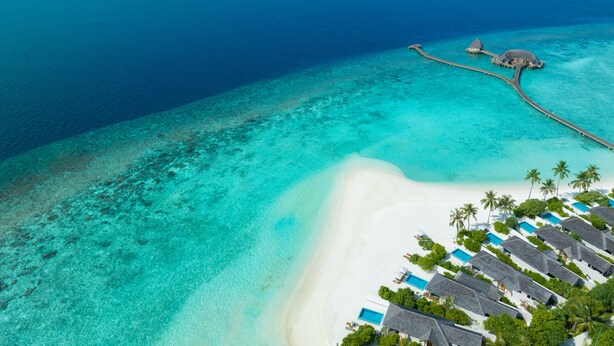 Vue aérienne d’un complexe hôtelier doté de bungalows sur pilotis, d’une plage de sable blanc et d’eaux turquoise cristallines.