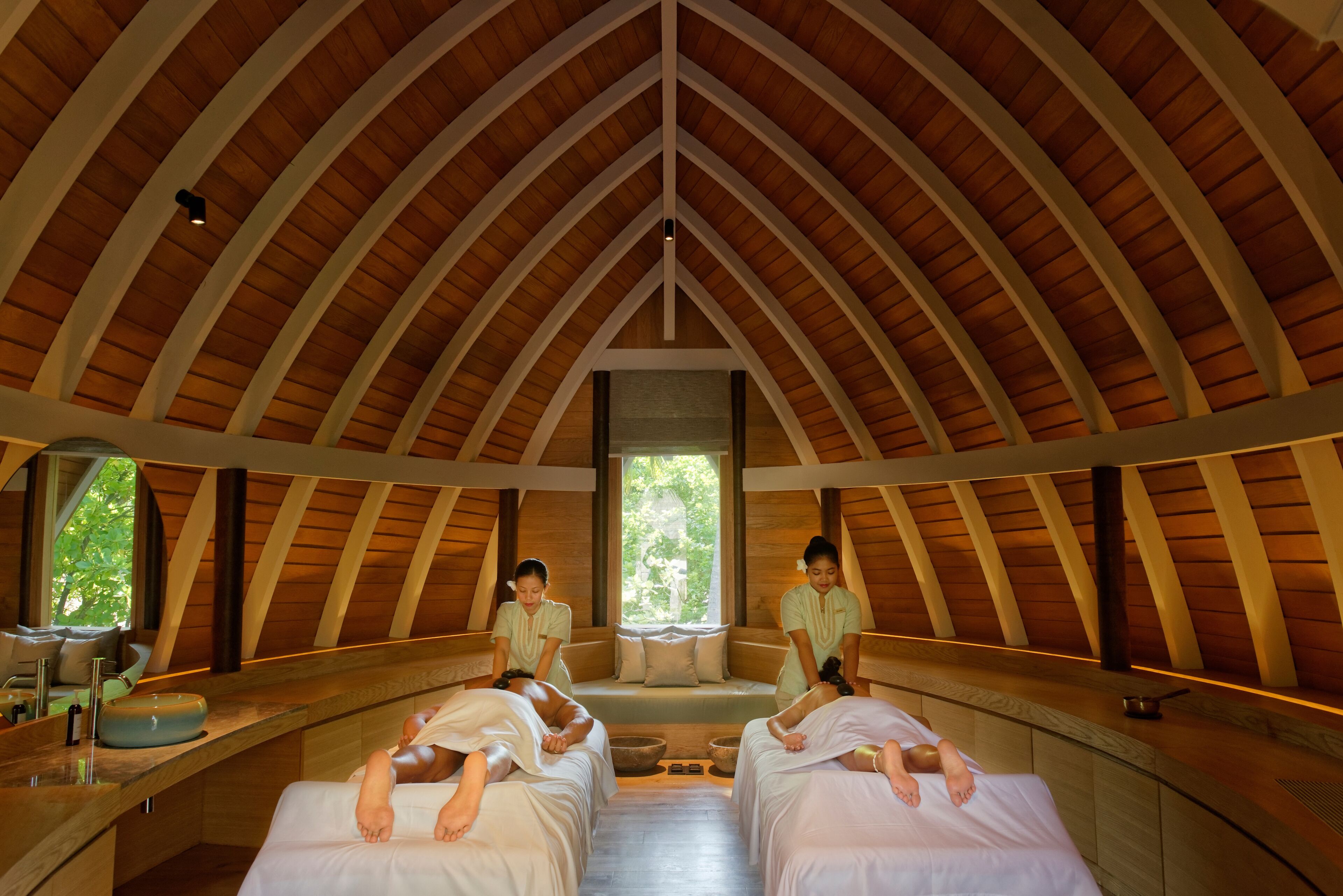 Een spa-ruimte met houten balken, twee massagetafels, een raam en een wastafel.