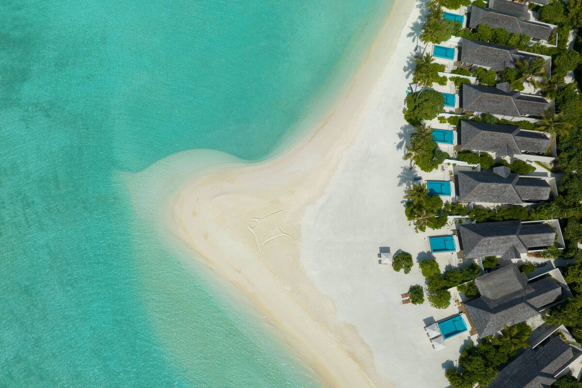 Ein Luftbild eines Strandresorts mit mehreren bungalowartigen Gebäuden, die jeweils einen Pool besitzen und entlang eines unberührten, weißen Sandstrandes mit klarem, türkisblauem Wasser liegen.