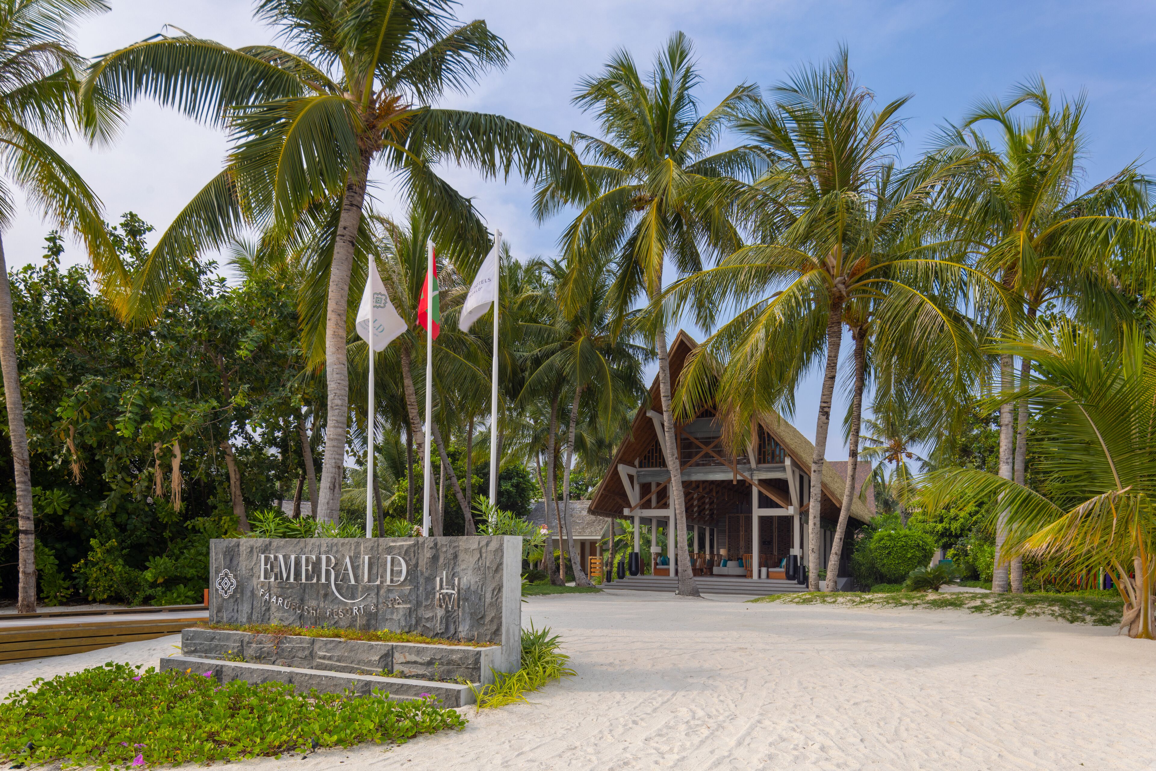 A resort entrance with a stone sign, palm trees, and a building with a thatched roof.