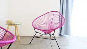 Two pink wire-framed chairs, a small round table with a basket and a book, and a grey curtain in a room with a plain white wall and grey flooring. - Hotel Mamá Carlota (Querétaro)