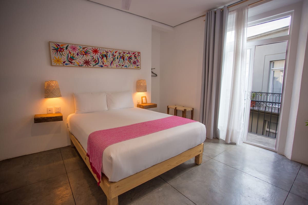 A bedroom with a bed, two bedside lamps, a nightstand, a colorful abstract painting, and a balcony with a railing.