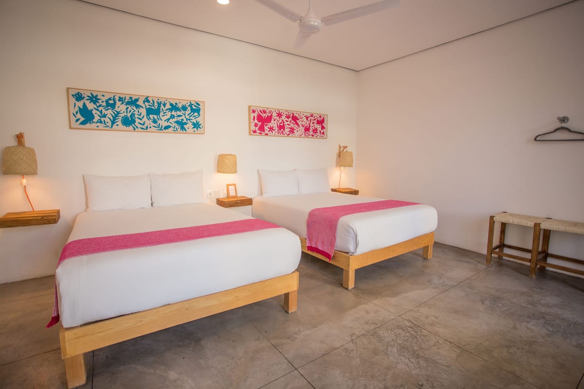 A hotel room with two beds, a ceiling fan, wall-mounted lamps, and a small table.