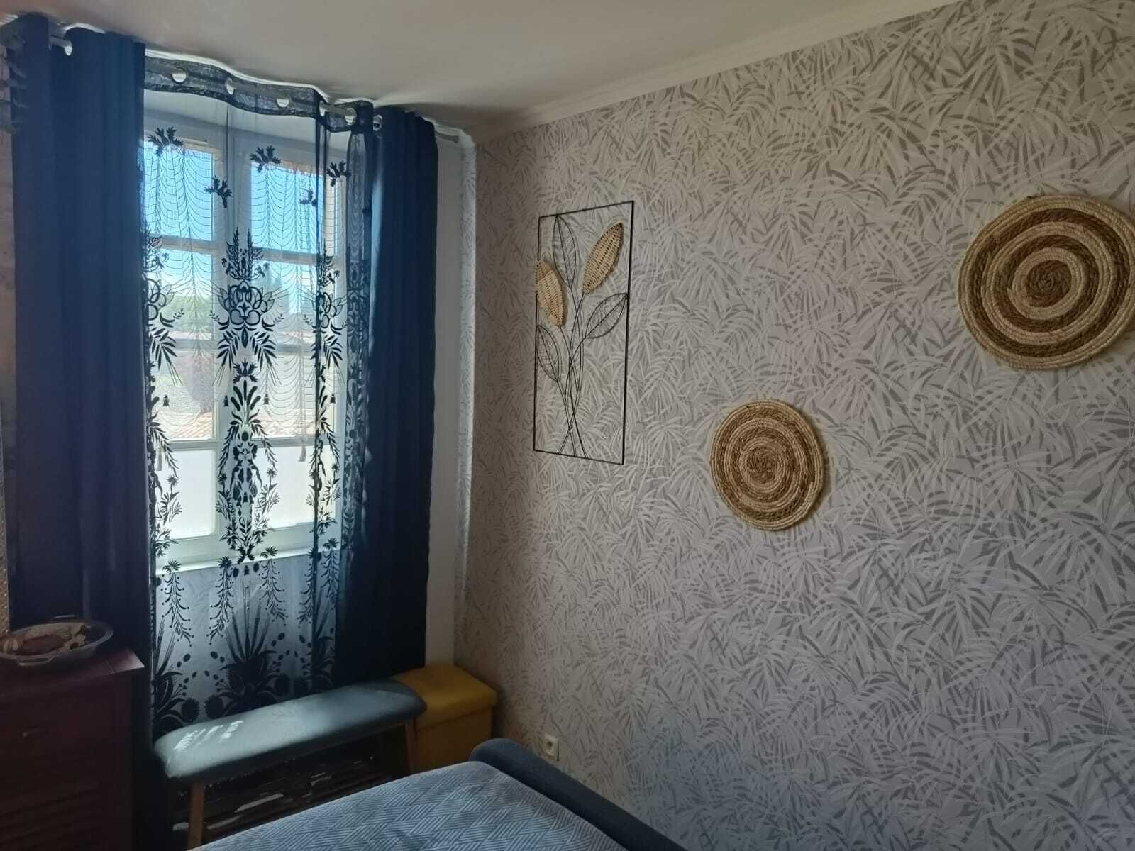 A room with a bed, a bench, a small stool, and a window with patterned curtains. There are two wall decorations and a textured wall.