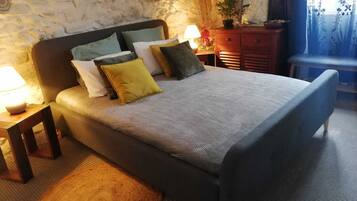 Una cama con colcha de estampado herringbone, una mesita de noche de madera con lámpara, una silla de mimbre y una pared de piedra.