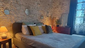 Een slaapkamer met een stenen muur, een bed met veel kussens, een nachtkastje met een lamp en een raam met gordijnen.