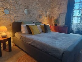 Un dormitorio con pared de piedra, una cama con muchas almohadas, una mesita de noche con lámpara y una ventana con cortinas.