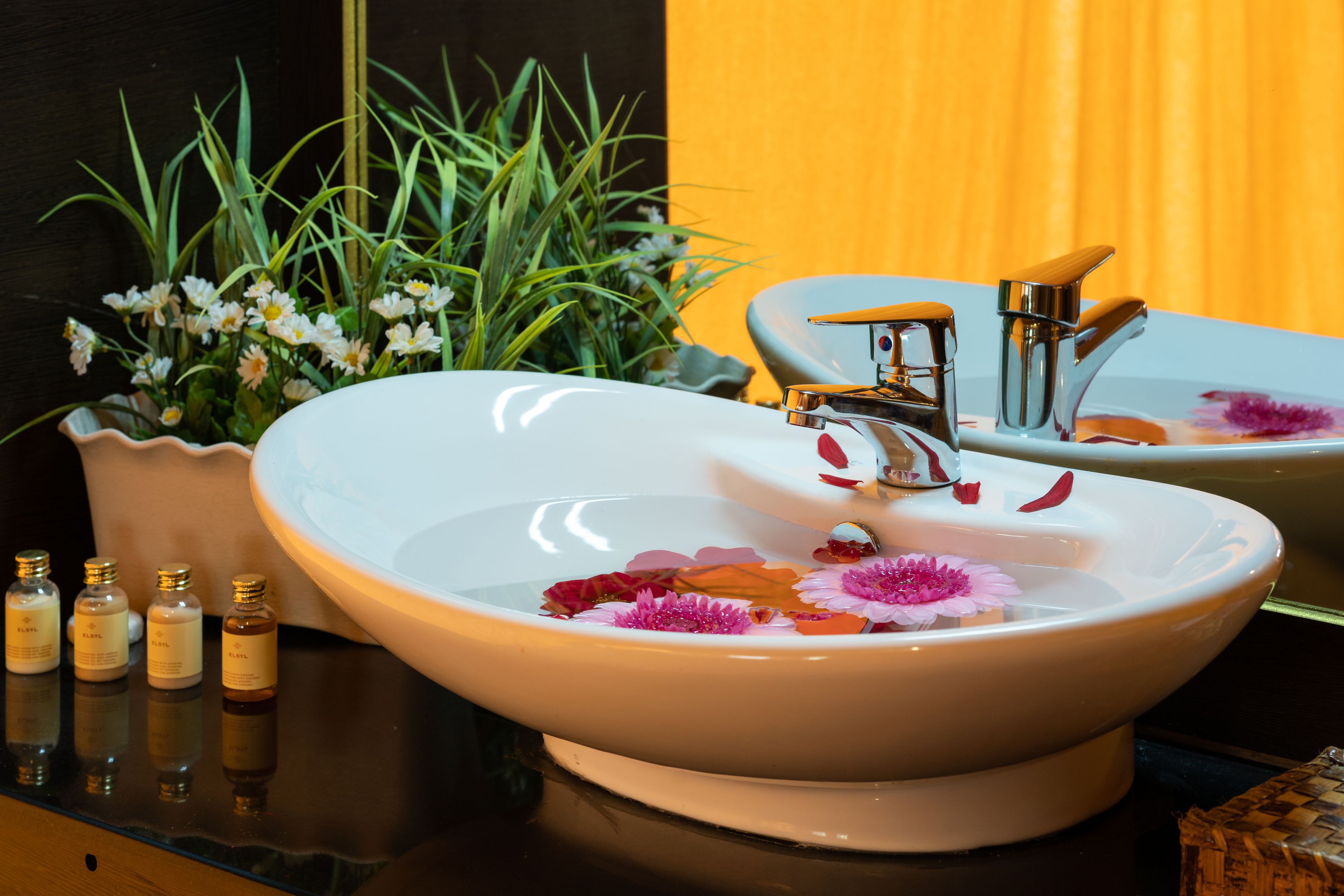Un baño con un lavamanos blanco, un espejo y una planta en maceta con flores blancas.