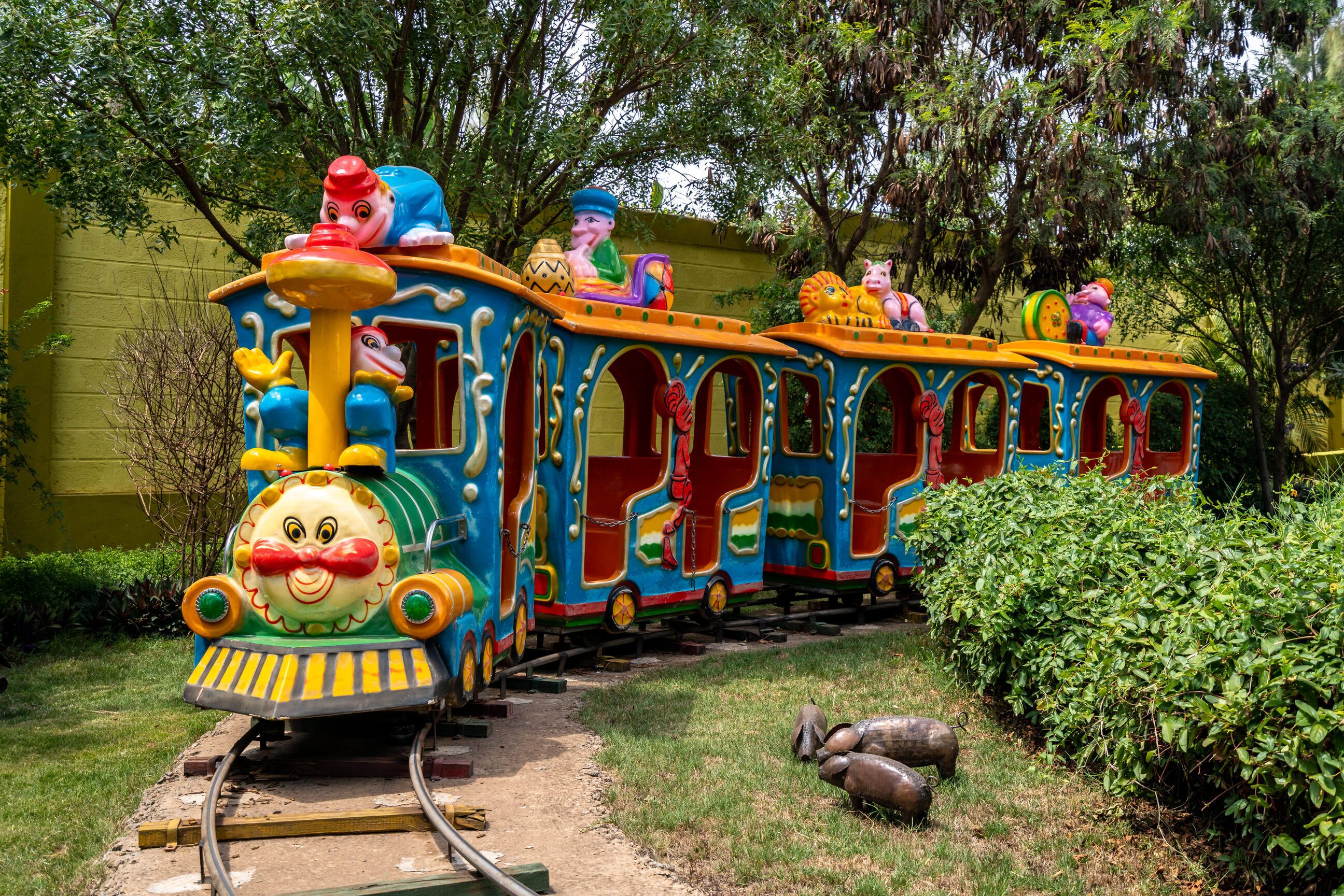 A colorful train with whimsical animal characters, set on tracks in a garden area with greenery and a yellow wall in the background.