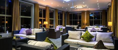 A well-lit indoor seating area with multiple sofas and chairs, large windows, and a chandelier.
