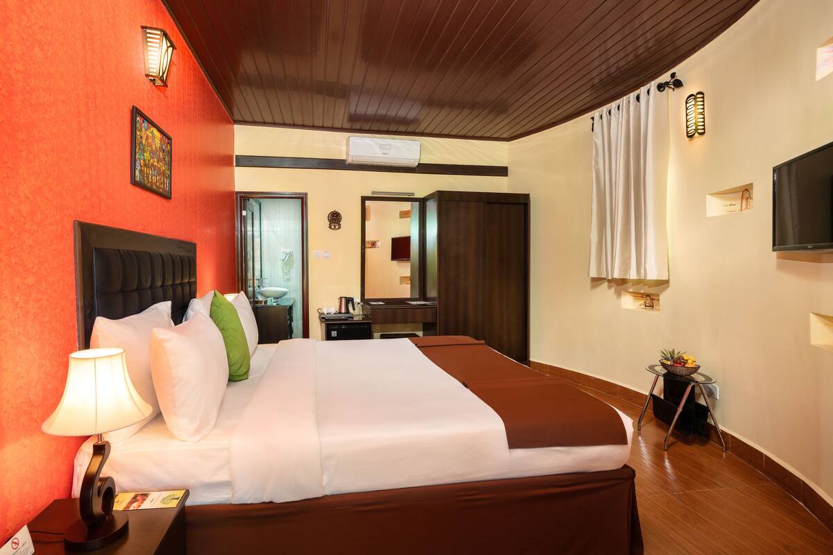 A hotel room with a large bed, a bedside table with a lamp, a TV, and a small table with fruit.