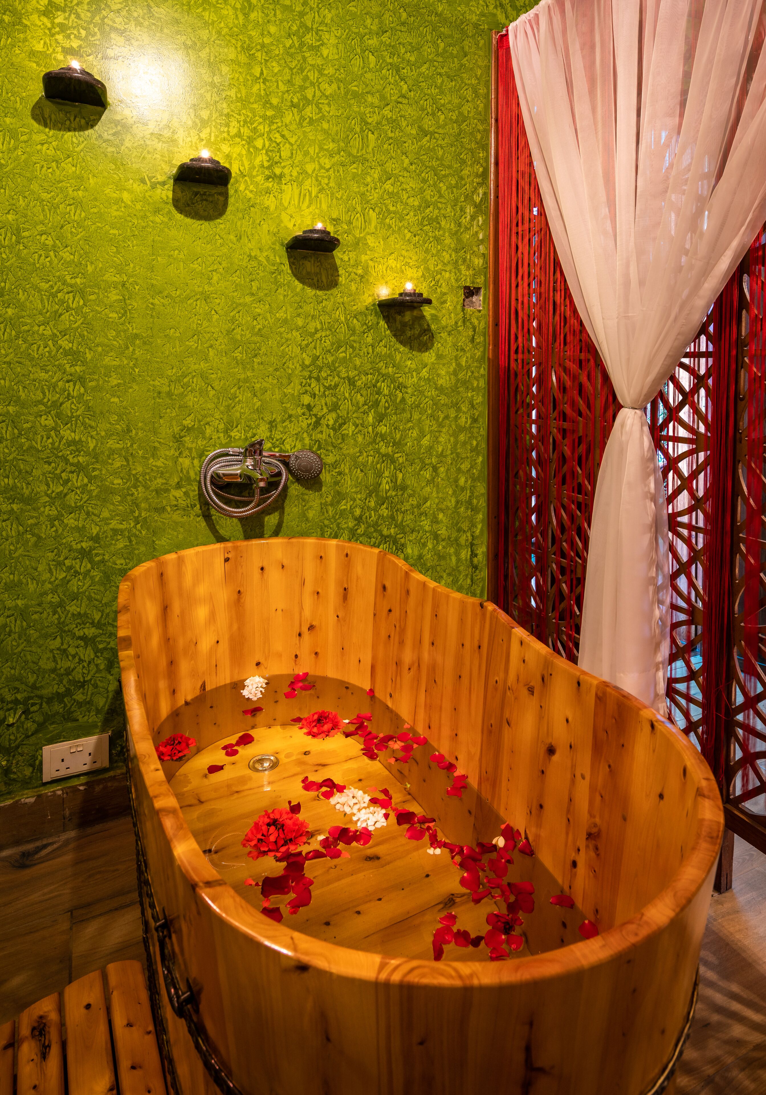 A wooden bathtub with rose petals, a green textured wall, and a red lattice partition.