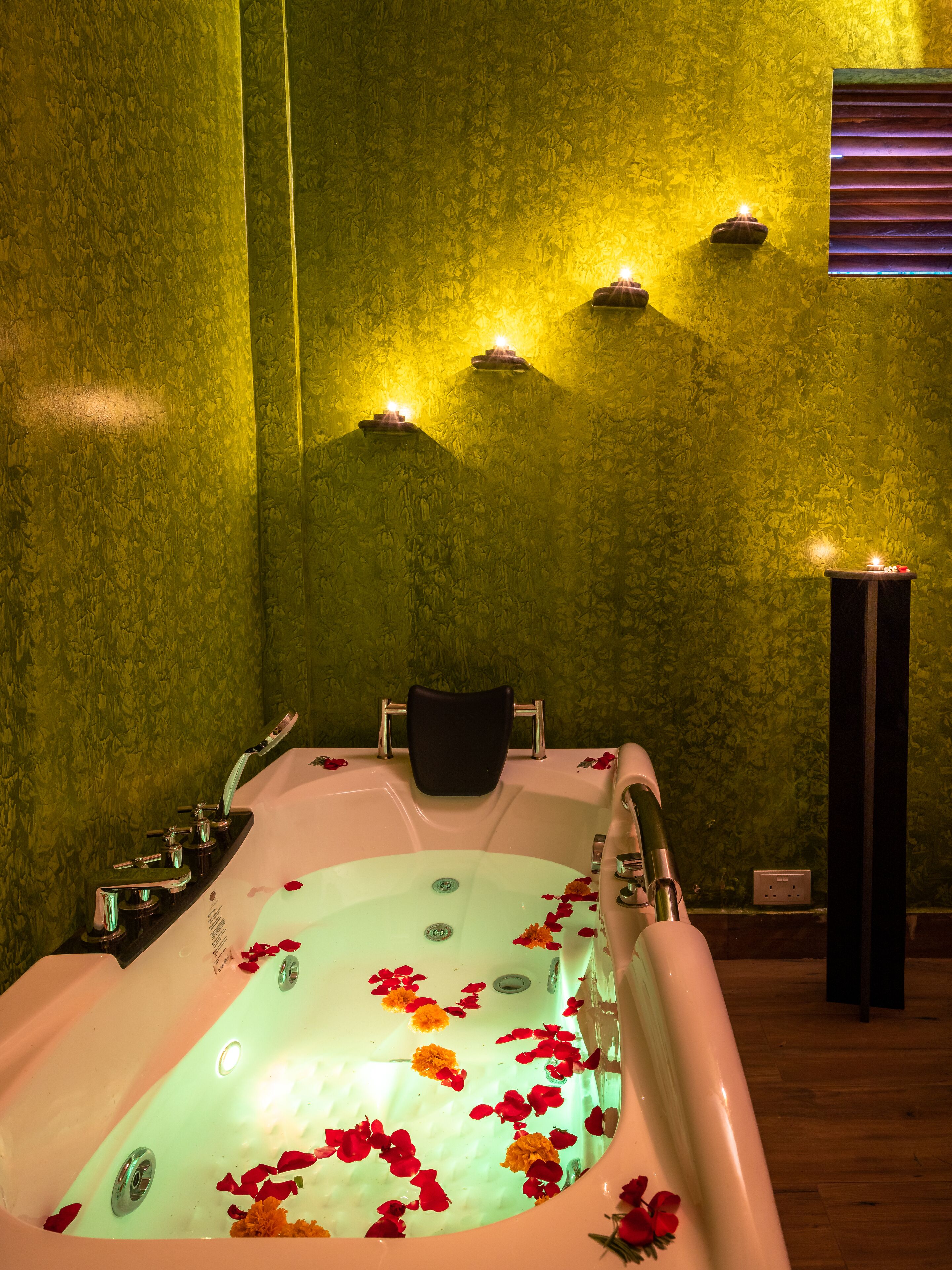 Une salle de spa avec une baignoire remplie d’eau et de pétales de rose, un fauteuil noir et des lampes fixées au mur.