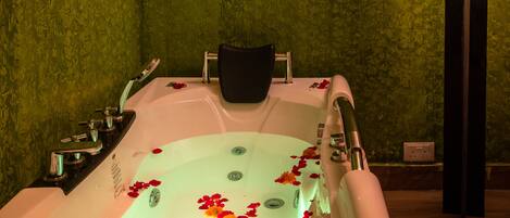 Un cuarto de spa con una bañera llena de agua y pétalos de rosa, una silla negra y luces fijadas a la pared.