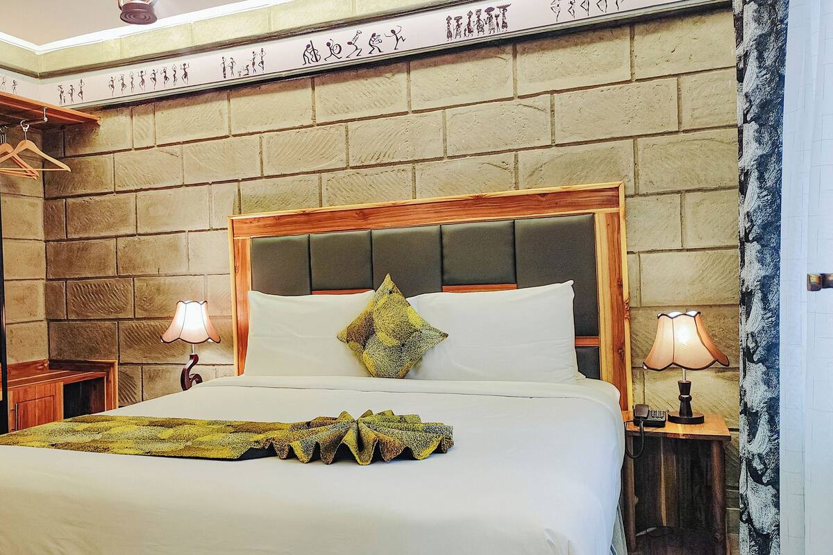 A hotel room with a bed, a wooden headboard, a bedside table with a lamp, a wall-mounted shelf with a radio, and a decorative wall piece.
