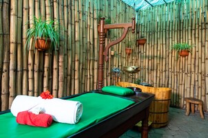 Un cuarto de spa con camilla de masaje, toallas verdes, un bidet de madera para pies y paredes de bambú.