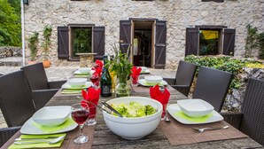 An outdoor dining table set with plates, bowls, cutlery, and wine glasses, with a stone building in the background. - Moulin d'Entre Les Roches (Puivert)