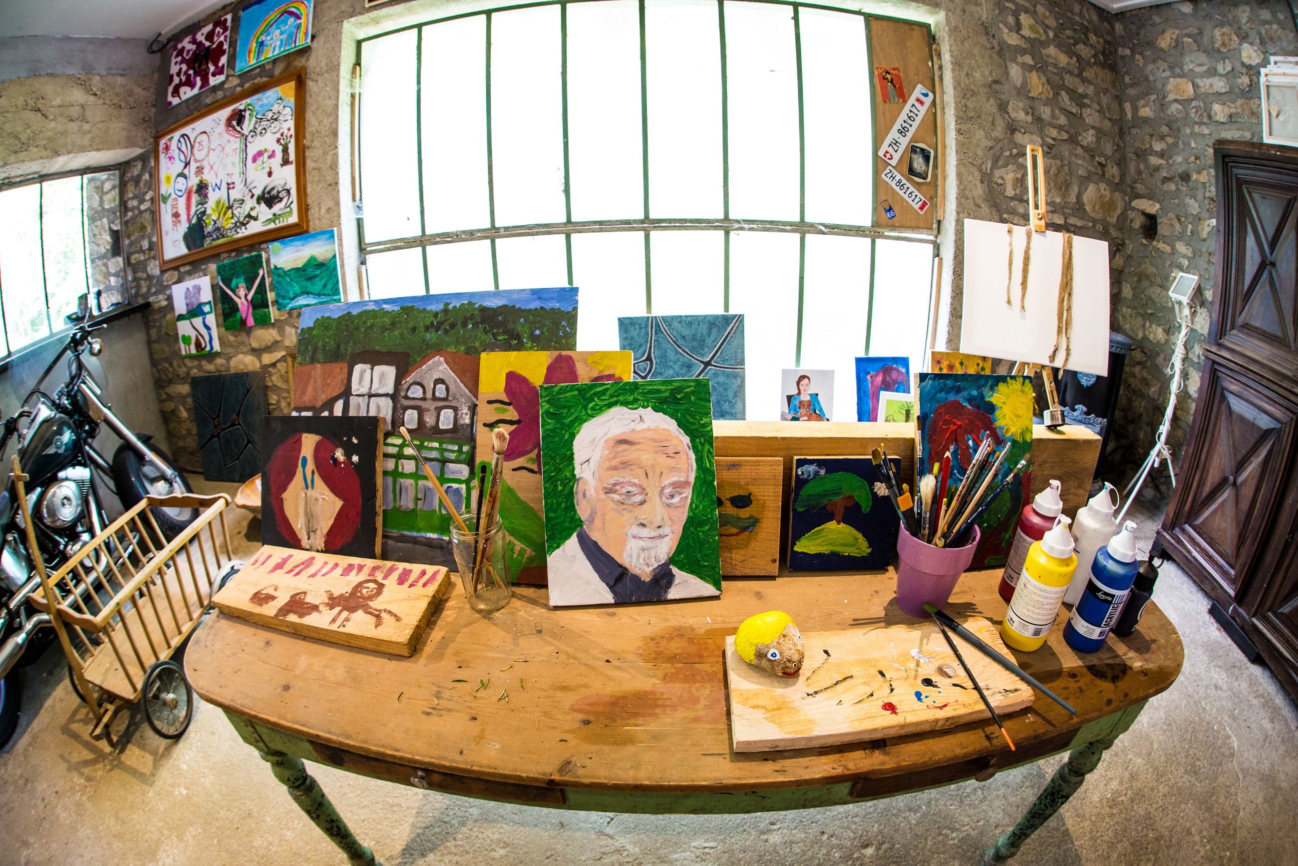 Une pièce où l’on trouve une table en bois sur laquelle sont exposées diverses peintures, un vélo et du matériel d’art.