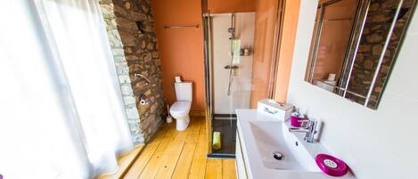 Une salle de bain avec un mur en pierre, un plancher en bois, un lavabo blanc et une douche en verre.