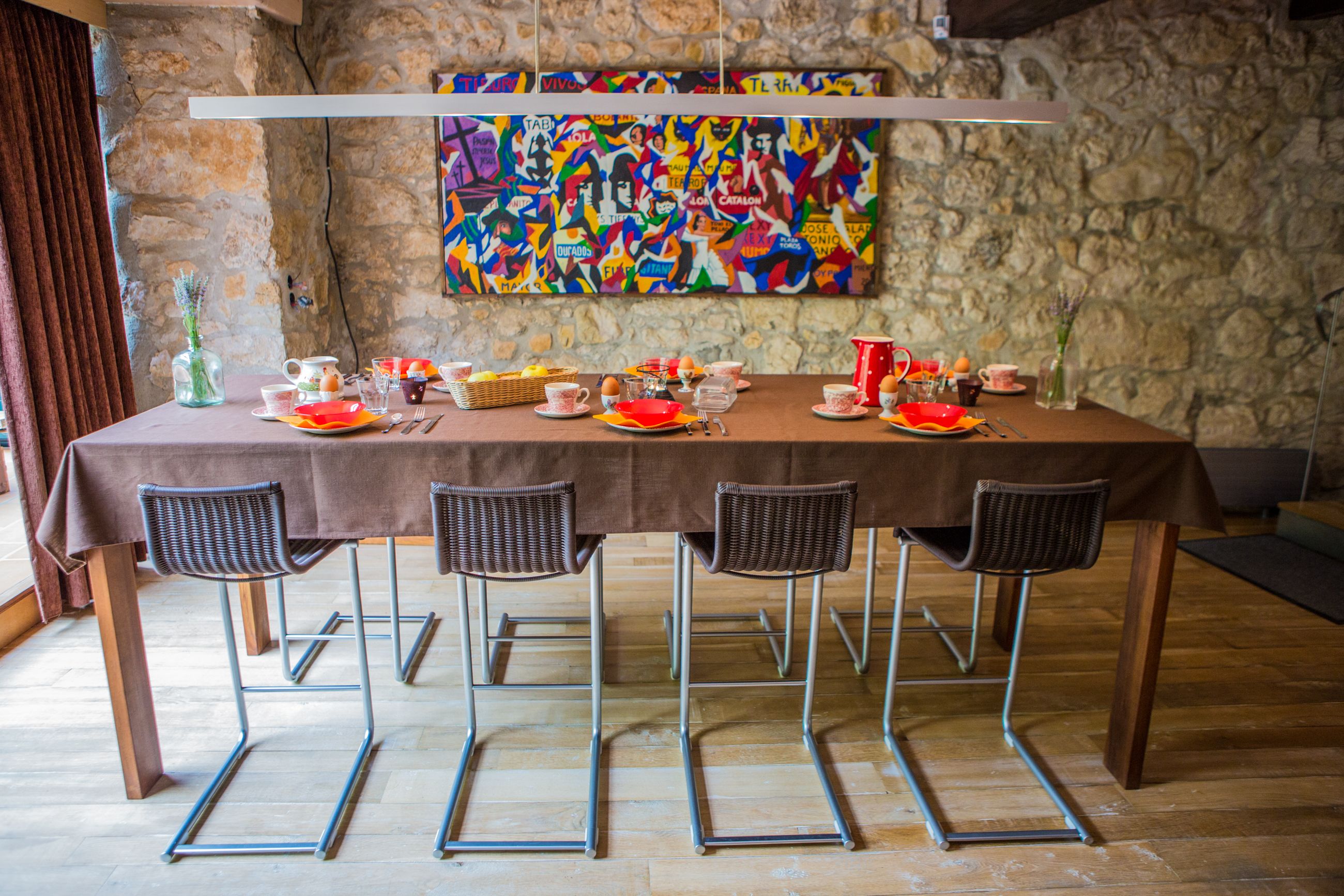 A dining table set with chairs, a colorful abstract painting on the wall, and a stone wall in the background.
