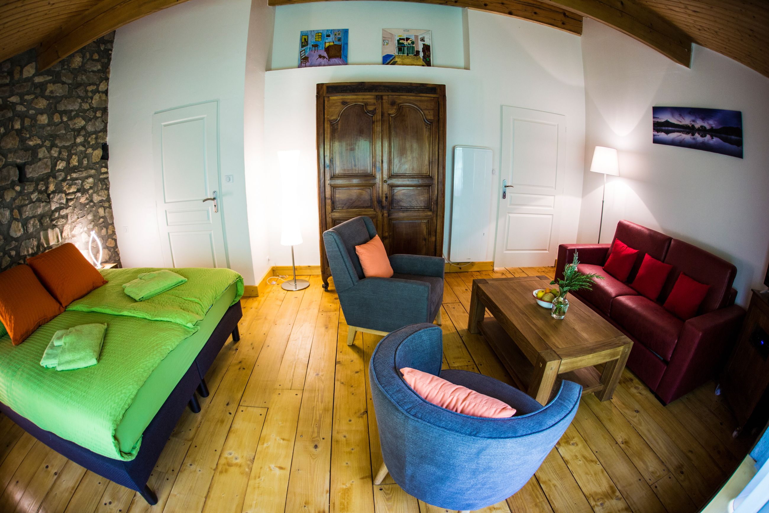 Una habitación con una cama, un sillón azul, un sofá rojo, una mesa de centro de madera y una pared de piedra.