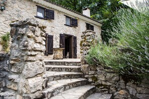 Un bâtiment en pierre, à l’aspect rustique, avec un escalier en pierre menant à une porte, et un chien qui se tient à l’entrée.