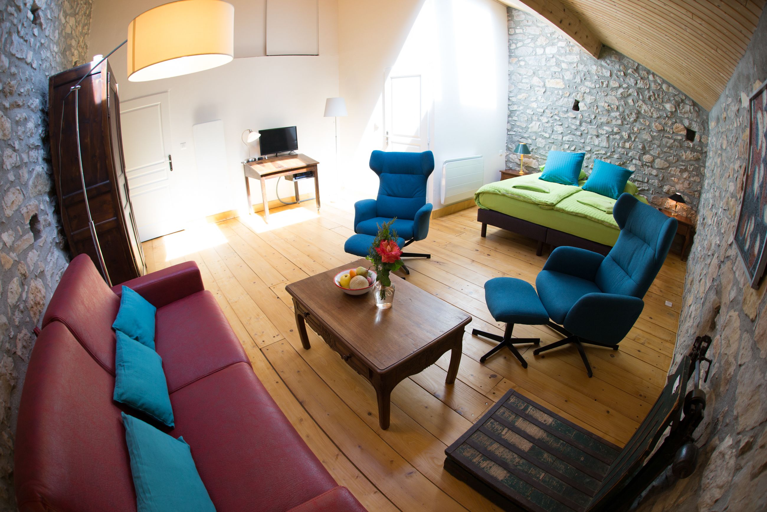 Una habitación acogedora con un sofá rojo, sillones azules, una mesa de centro de madera con un jarrón y una cama con ropa de cama verde.