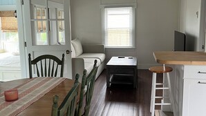 Et lille køkken med spisebord, stole, et træsofabord, en loftsvifter og et vindue med persienner.