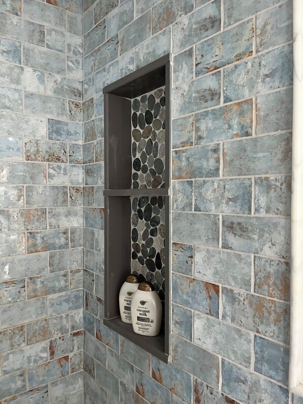 A bathroom with a blue and gray brick wall, a shelf with bottles of coconut milk shampoo, and a small window. - Classic Beach Cottage, Steps to Ocean Break. (Narragansett)