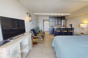 A hotel room with a TV, a bed, a dining area with chairs, a kitchen, and a lamp.