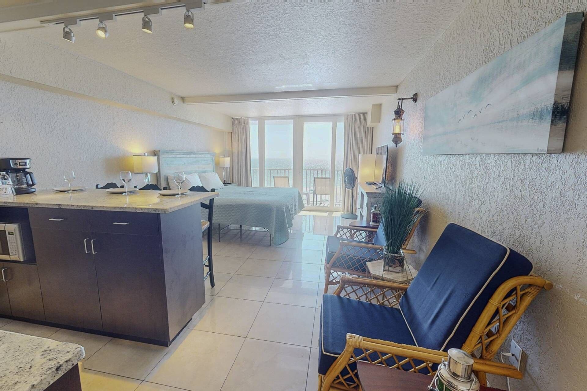 A hotel room with a bed, a dining area with a table and chairs, a kitchen area, and a balcony with a view.