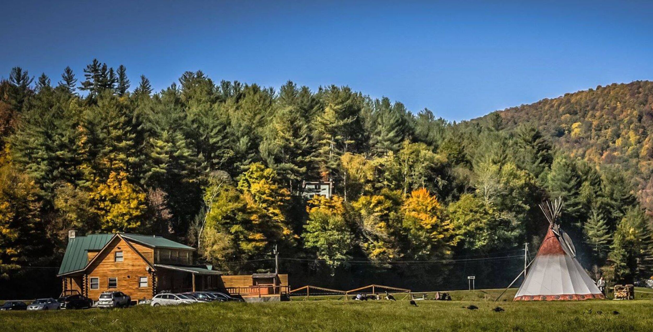 A lodge with a green roof, a teepee, and a forested hillside in the background.