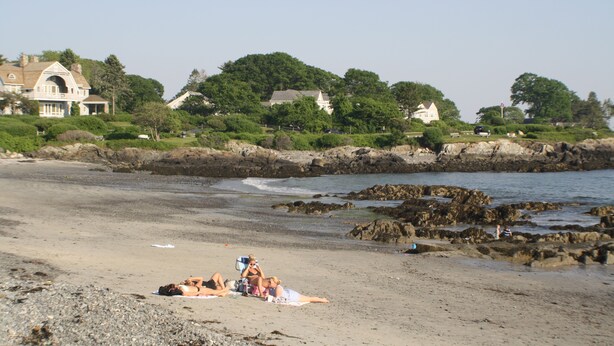Una playa con algunas personas tomando el sol, una costa rocosa y casas al fondo.