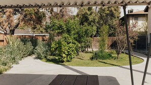 A covered patio with a wooden table, surrounded by a garden with various plants and trees.