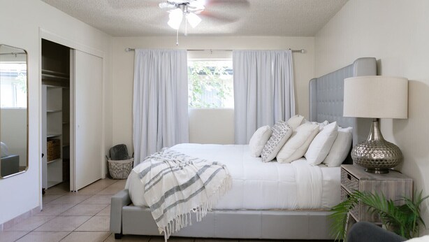 A bedroom with a bed, a bedside lamp, a window with curtains, a ceiling fan, and a view of the outdoors.