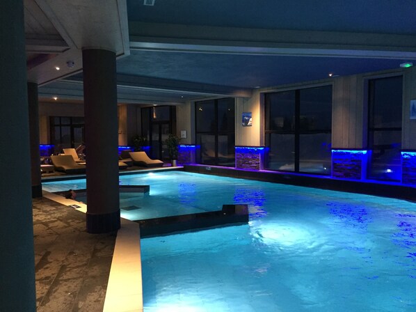 Una piscina interior con vista a la ciudad de noche, rodeada de sillones de descanso y un muro de piedra con iluminación azul.