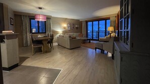 Habitació d'hotel moderna amb zona de menjador, saló amb sofà i butaques, balcó amb vista a les muntanyes i finestra gran amb cortines.