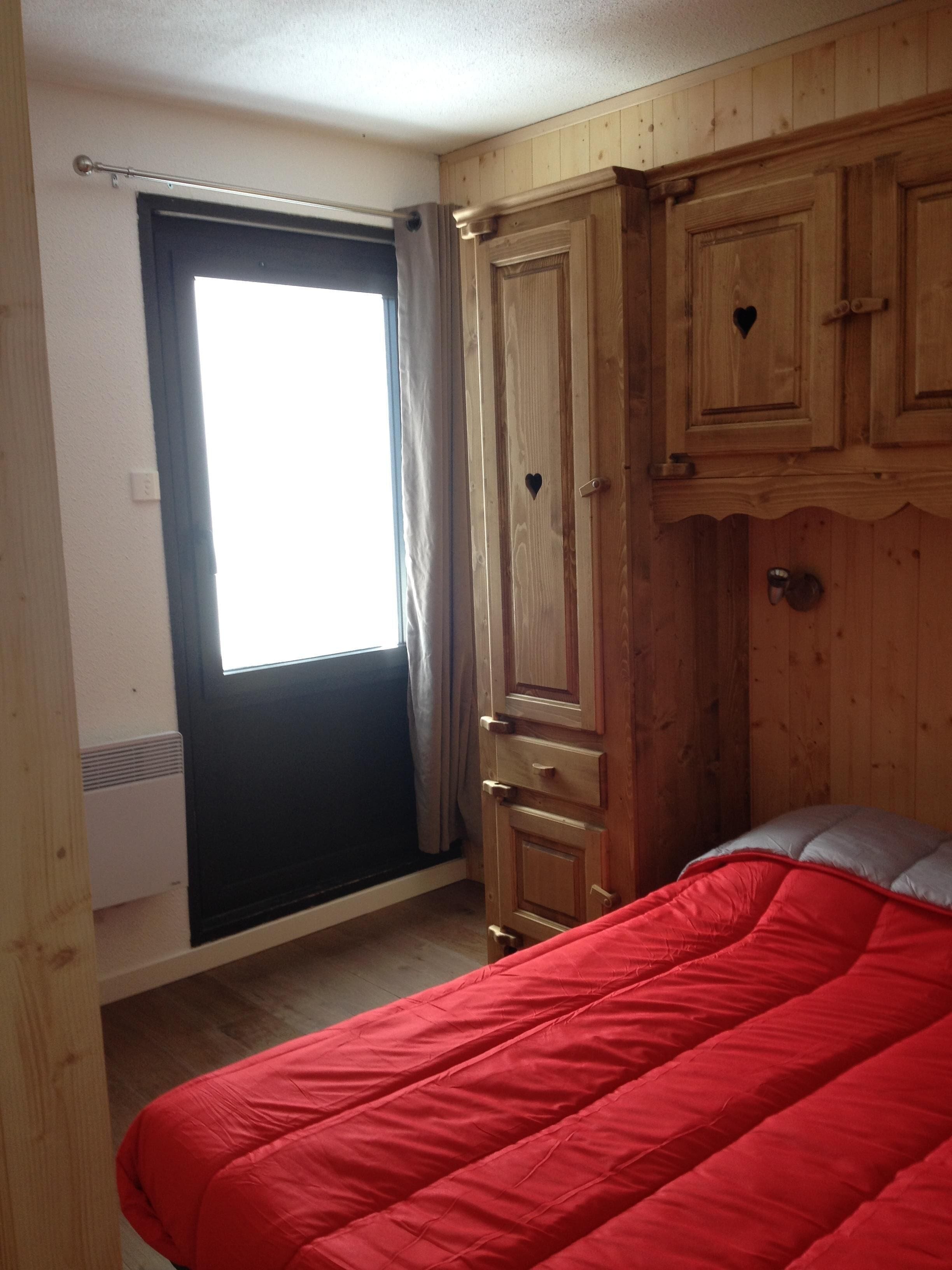 Une chambre à coucher avec un lit rouge, une armoire en bois et une fenêtre avec des rideaux.
