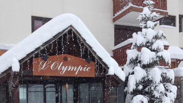 Entrada de un hotel con un letrero que dice 'L'Olympic', rodeada de árboles cubiertos de nieve y suelo nevado.
