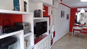 Een moderne woonkamer met een flatscreen televisie, een boekenkast, een eethoek met een rode tafel en stoelen, en een aquarium.