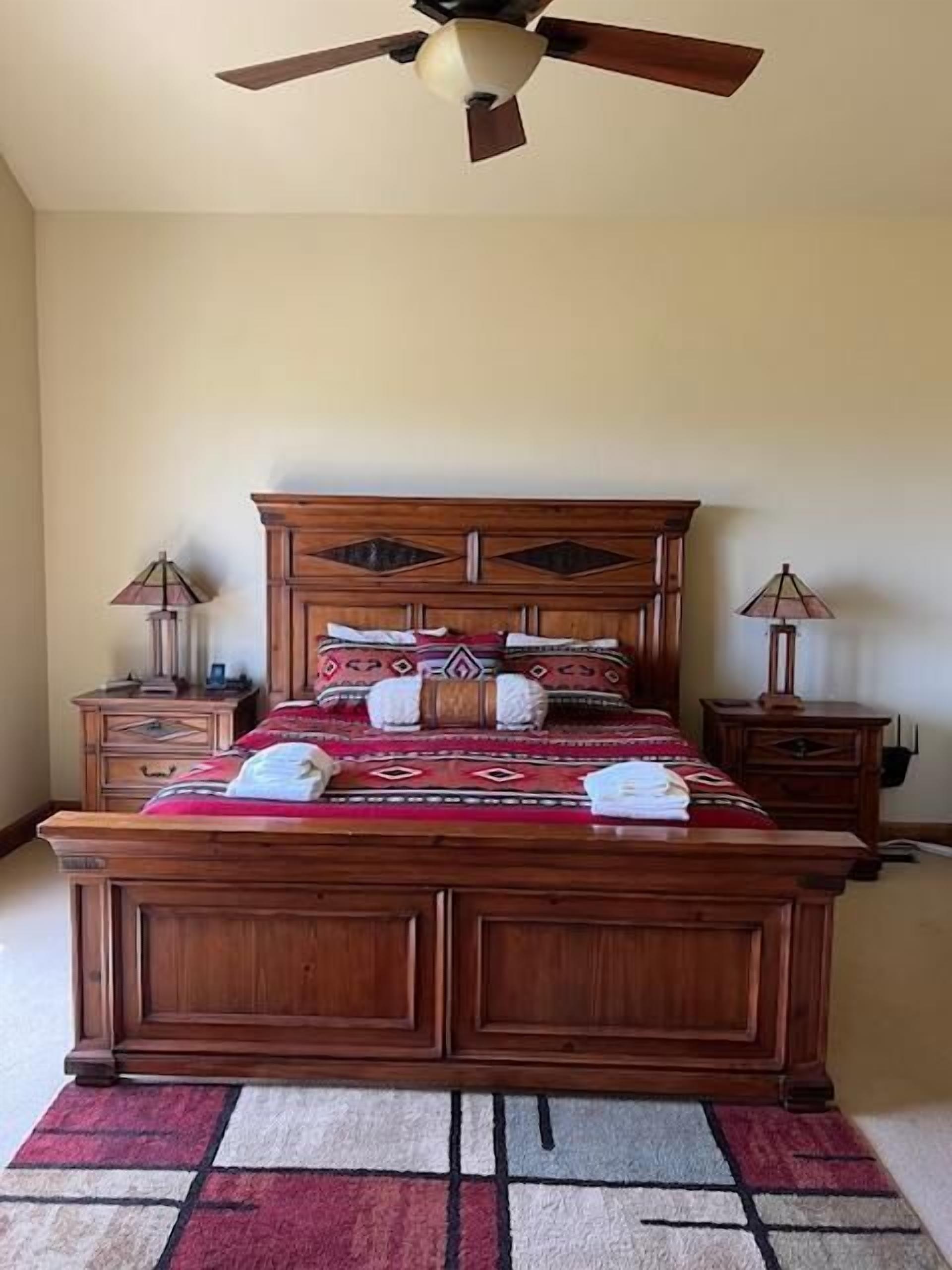A wooden four-poster bed with a red and white patterned bedspread, two bedside tables with lamps, and a ceiling fan.