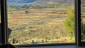 Une vue depuis une fenêtre donnant sur une vallée, avec des montagnes au loin, des champs et un petit plan d’eau.