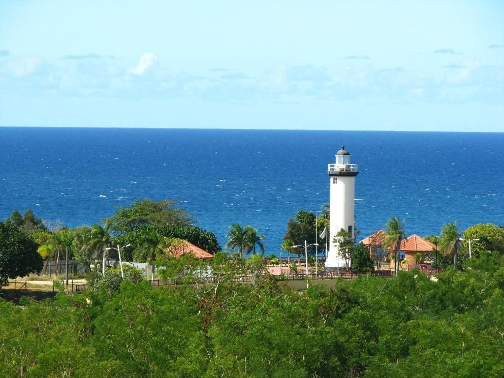 Ein Leuchtturm mit freiem Blick auf den Ozean und tropischer Umgebung, mit Palmen und Gebäuden im Hintergrund.