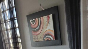 A small dining area with a table and two chairs, a framed abstract painting on the wall, and a window with curtains. - Boricua Apartments (Rincon)