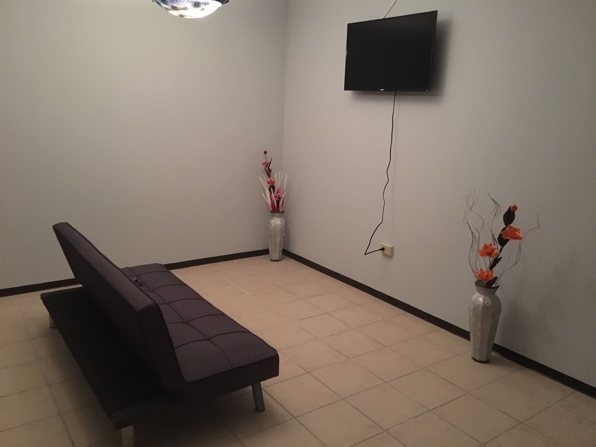 A room with a dark-colored sofa, two white vases with flowers, a flat-screen TV mounted on the wall, and a tiled floor.