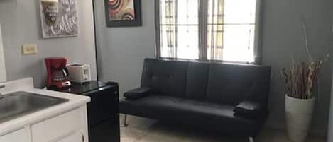 A compact kitchen with a sink, a coffee maker, and a refrigerator. There is a black sofa against the wall and a window with blinds.