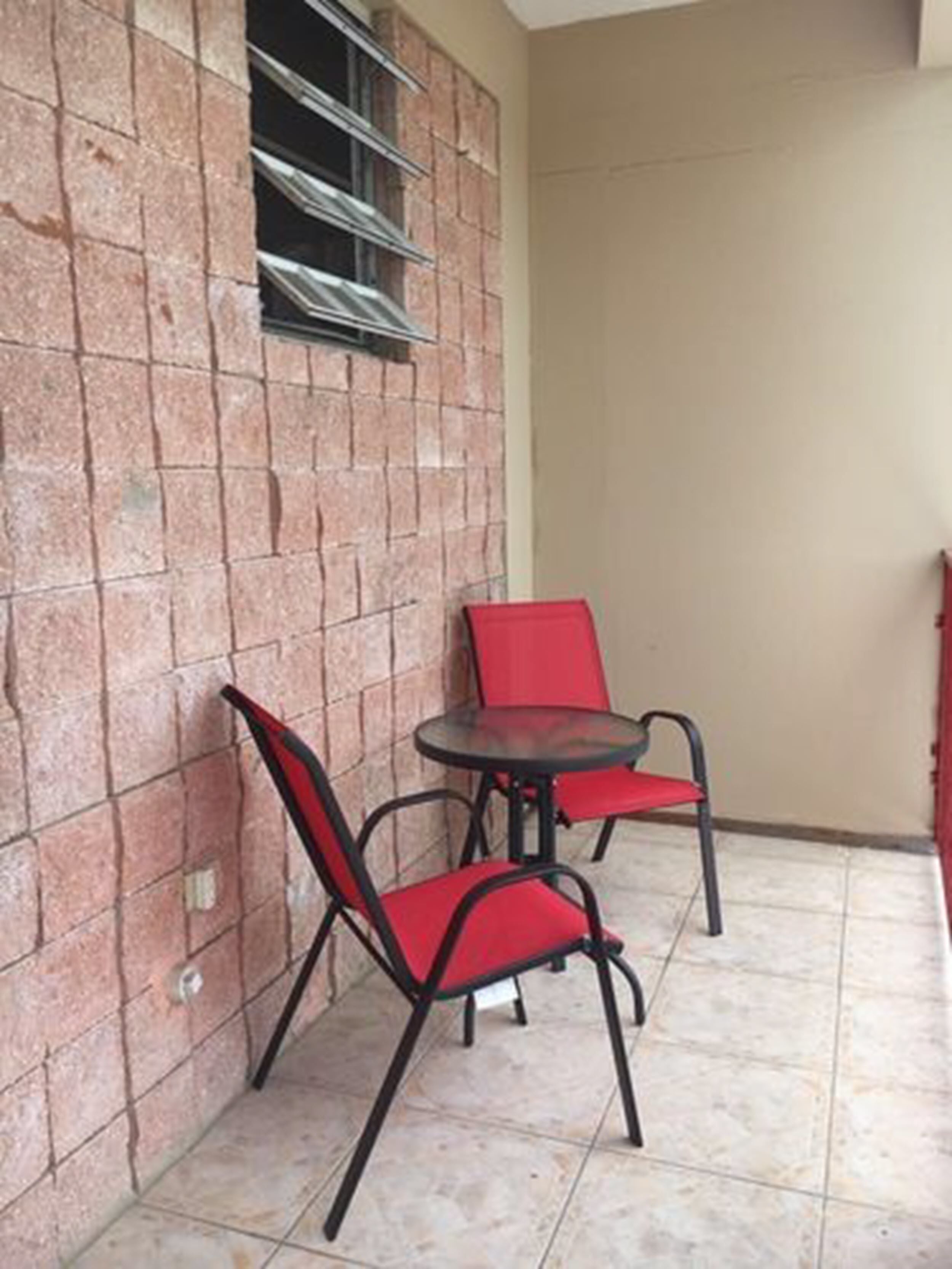 A small outdoor seating area with two red chairs and a table, adjacent to a brick wall with a window.