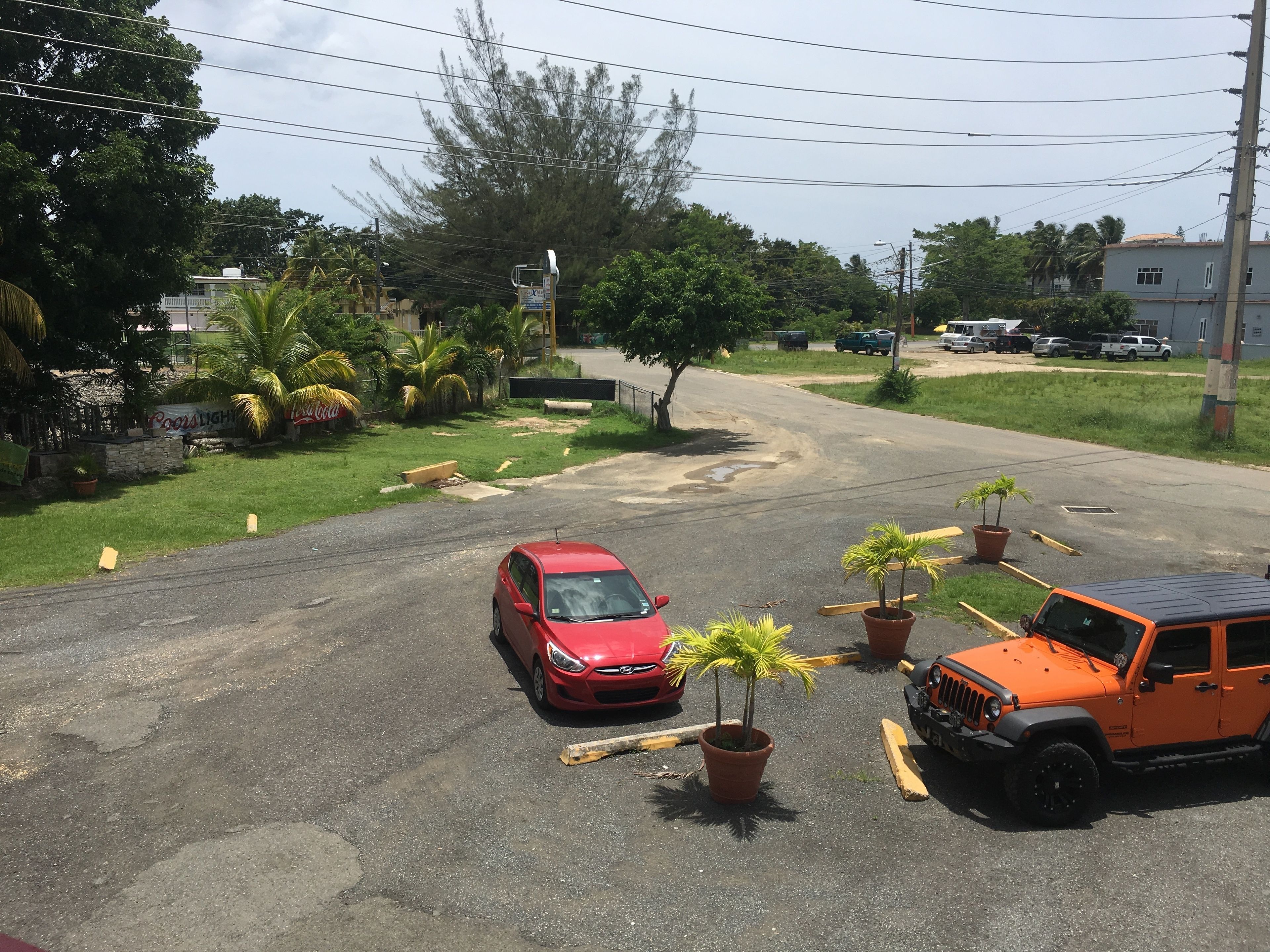 Un parking avec une voiture rouge et un VUS orange. On y trouve des plantes en pot et un banc.