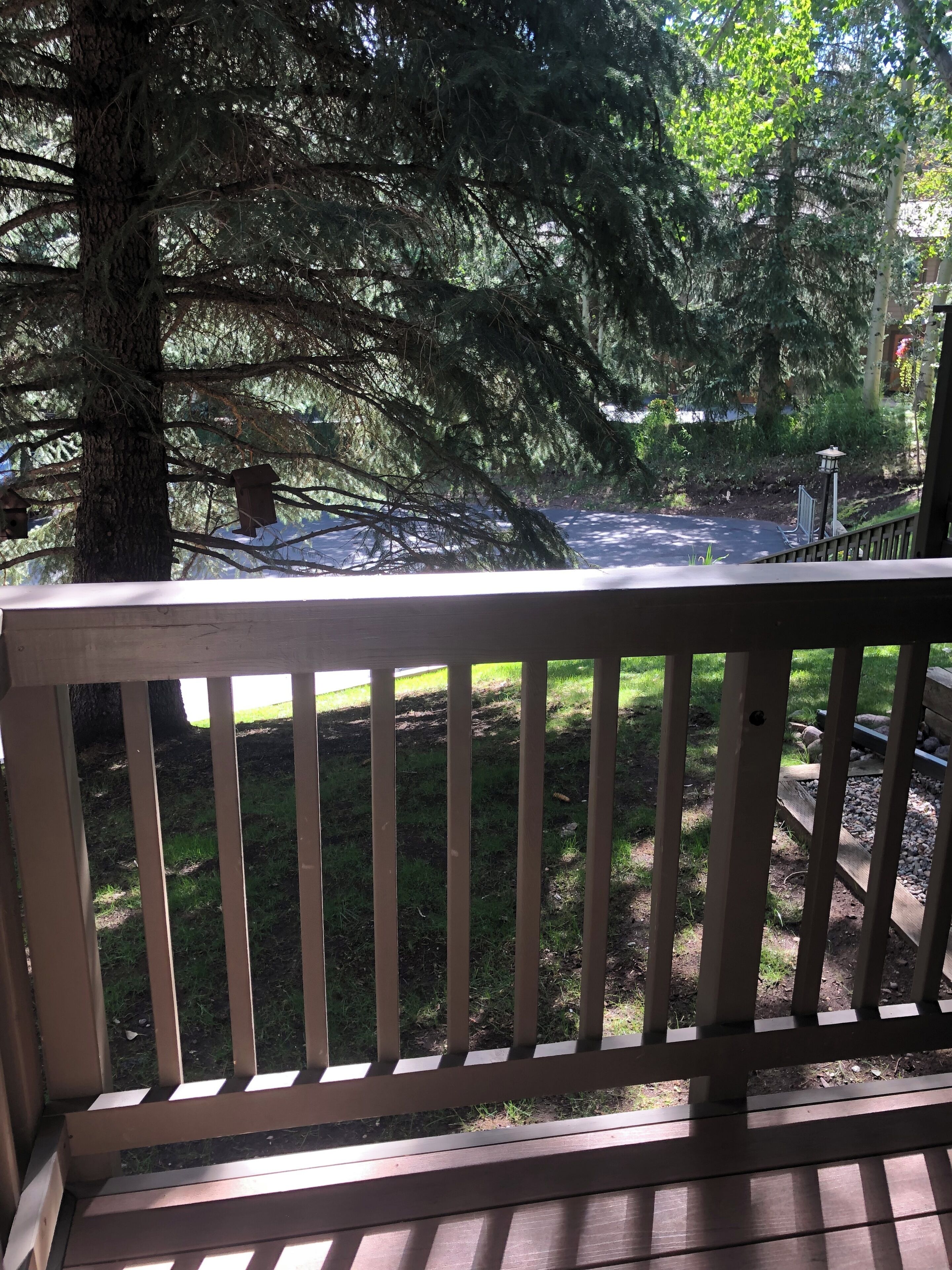 Une terrasse en bois dotée d’une rambarde, qui donne sur un jardin arboré et une zone dallée.