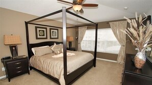 Una cama con dosel, un ventilador de techo, mesitas de noche con lámparas y un florero con plantas secas sobre un aparador.